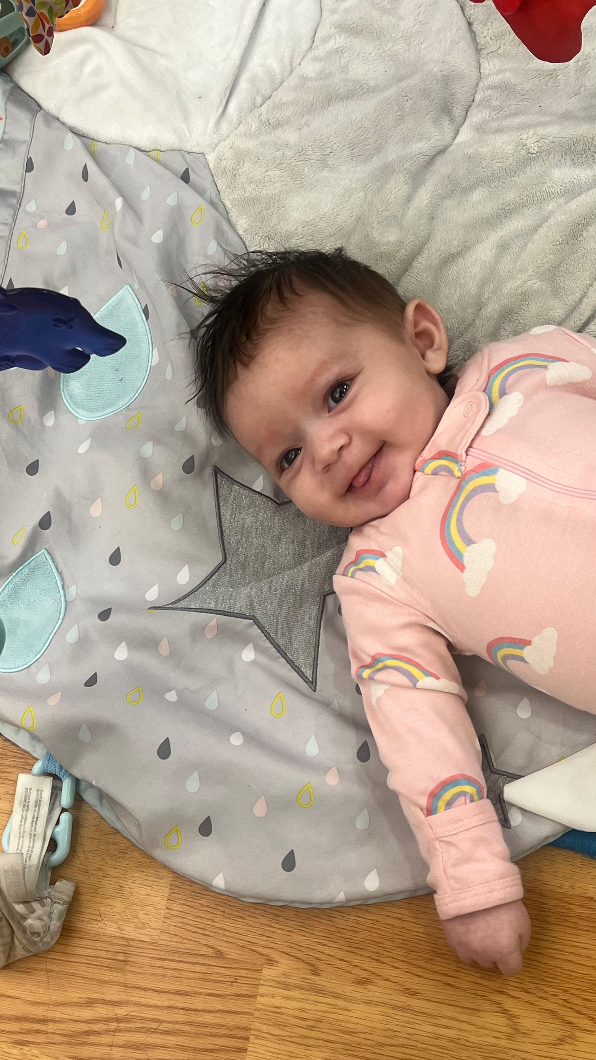 A smiling baby in a pink rainbow-patterned onesie lying on a gray star-themed play mat.
