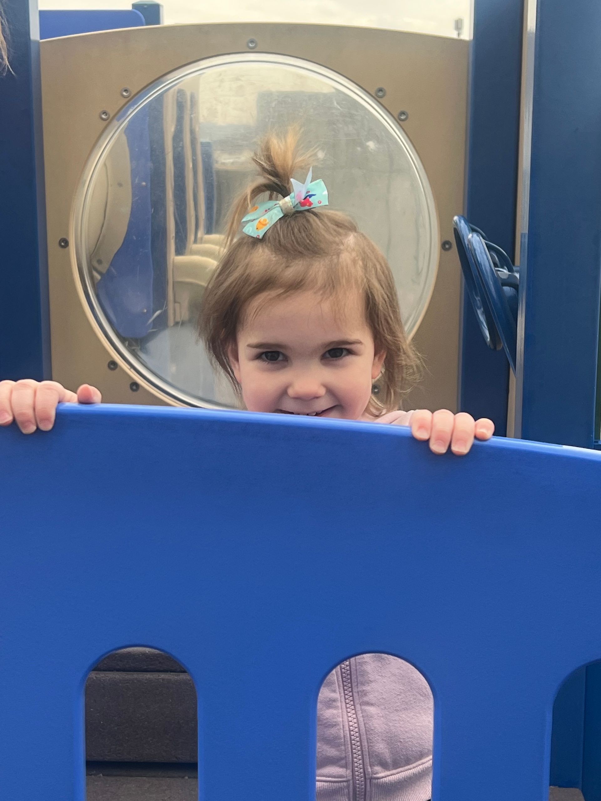 A young child with a hair bow peers over a blue plastic playground wall, smiling towards the camera.