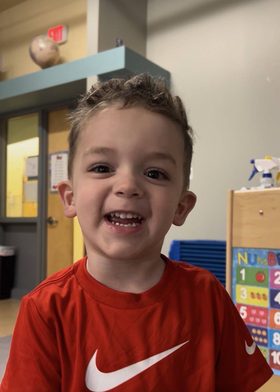 A child with curly hair in a red Nike shirt smiling widely in a classroom setting with a number chart in the background.