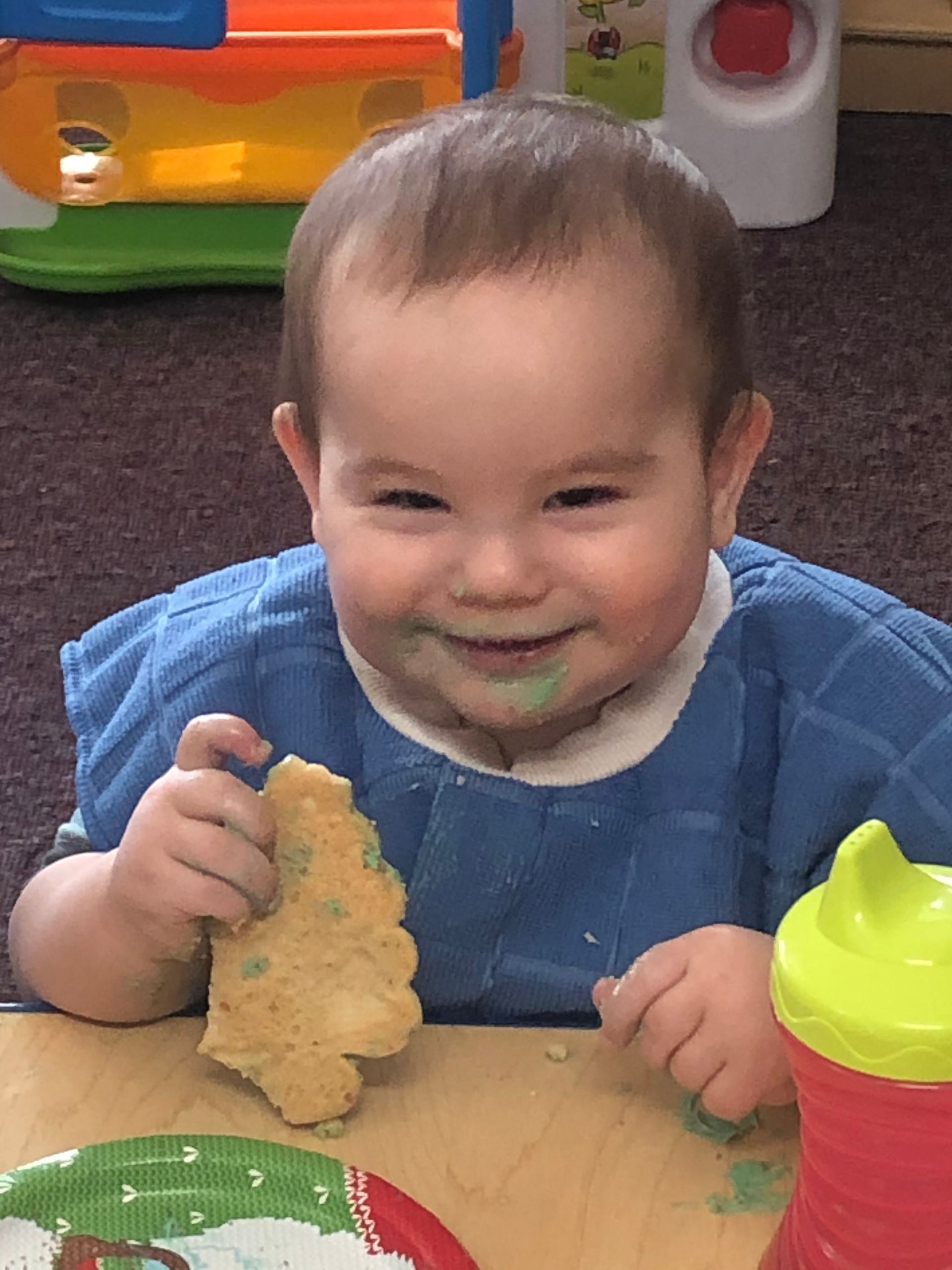 A smiling child in a blue bib holds a cookie at a table next to a green and red cup.