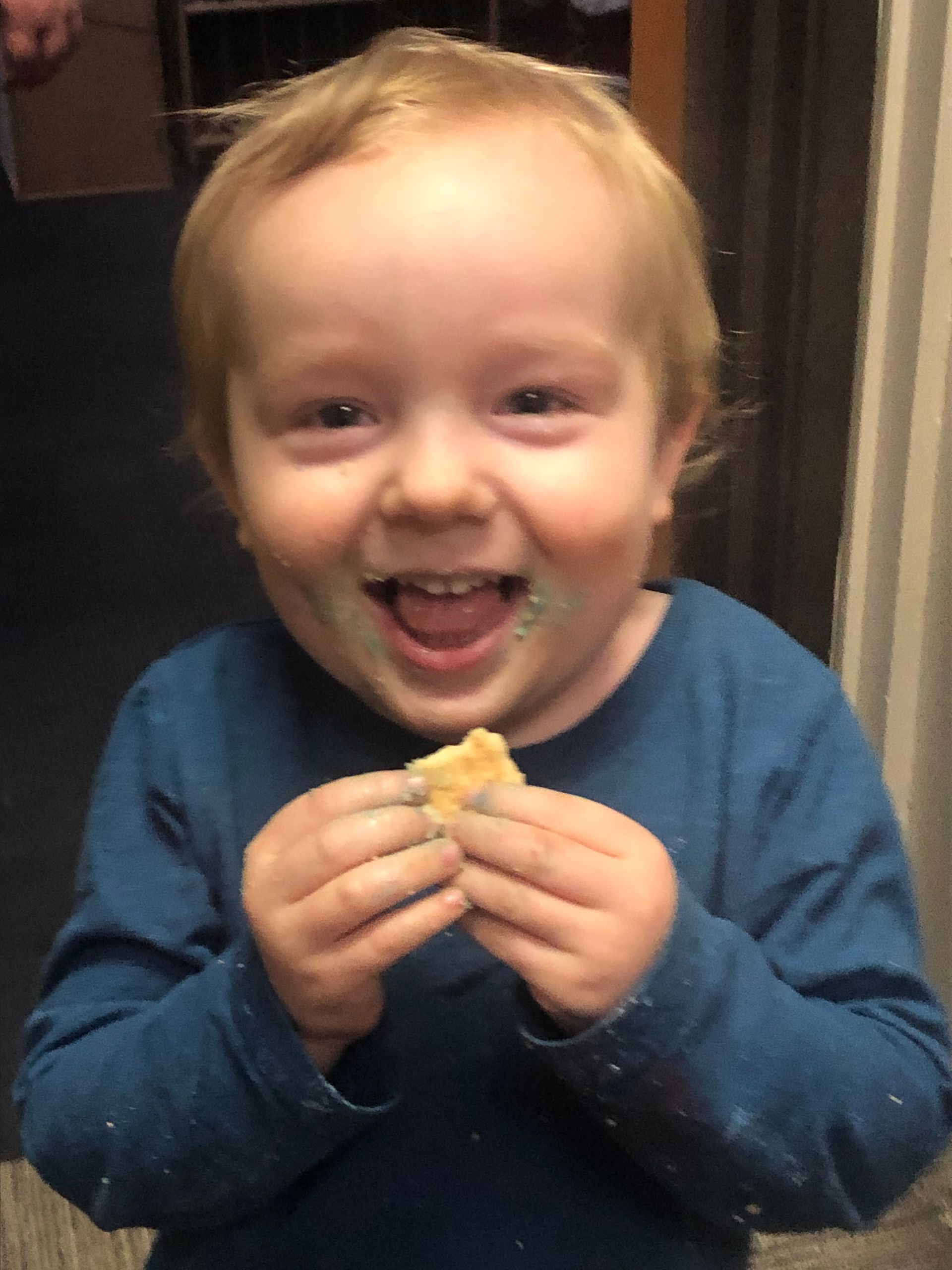A smiling child with blonde hair wearing a blue long-sleeved shirt, holding a cracker with residue on their hands and face.