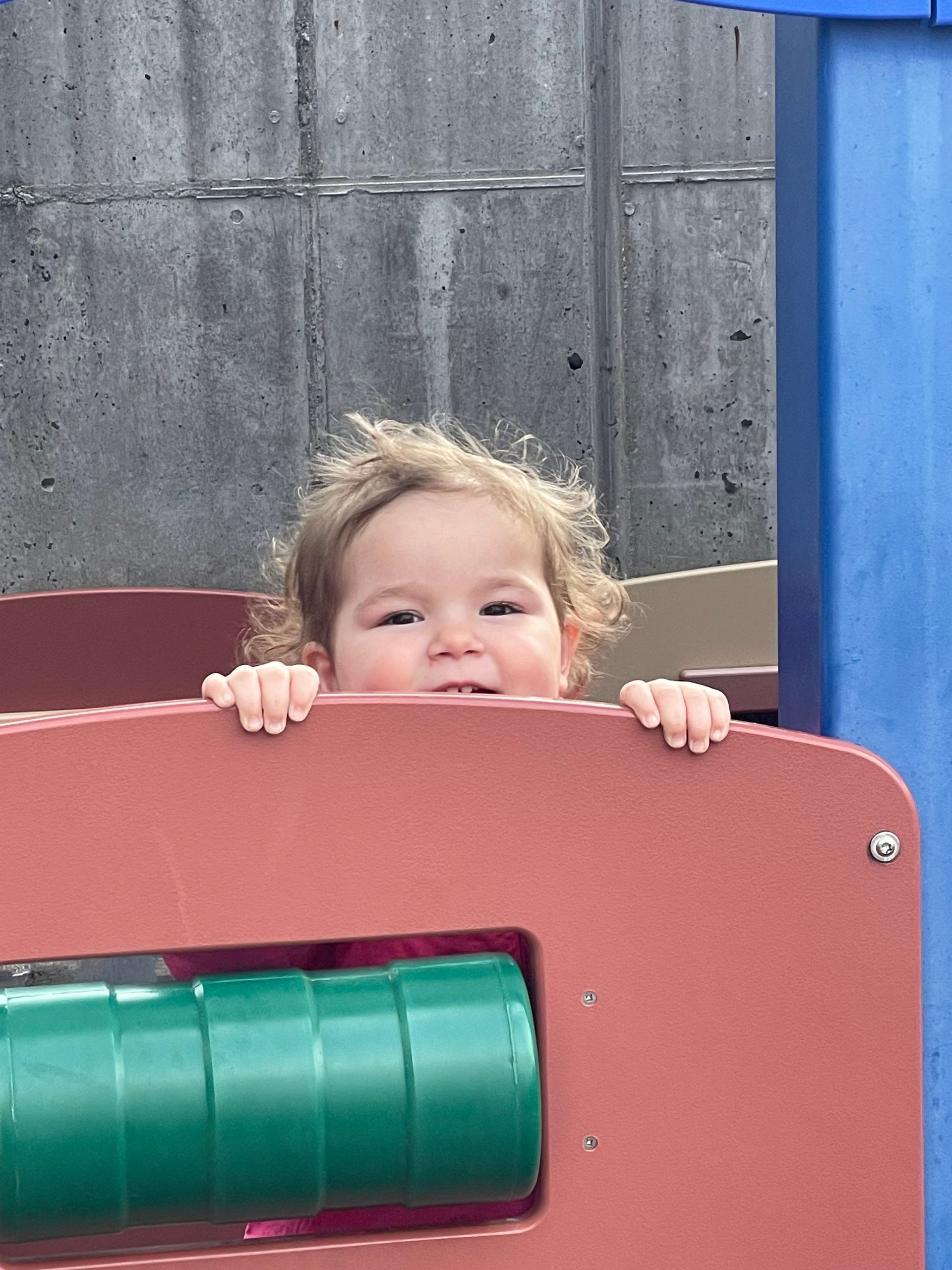 A toddler peeking over the top of a red plastic play structure with a green horizontal bar below.