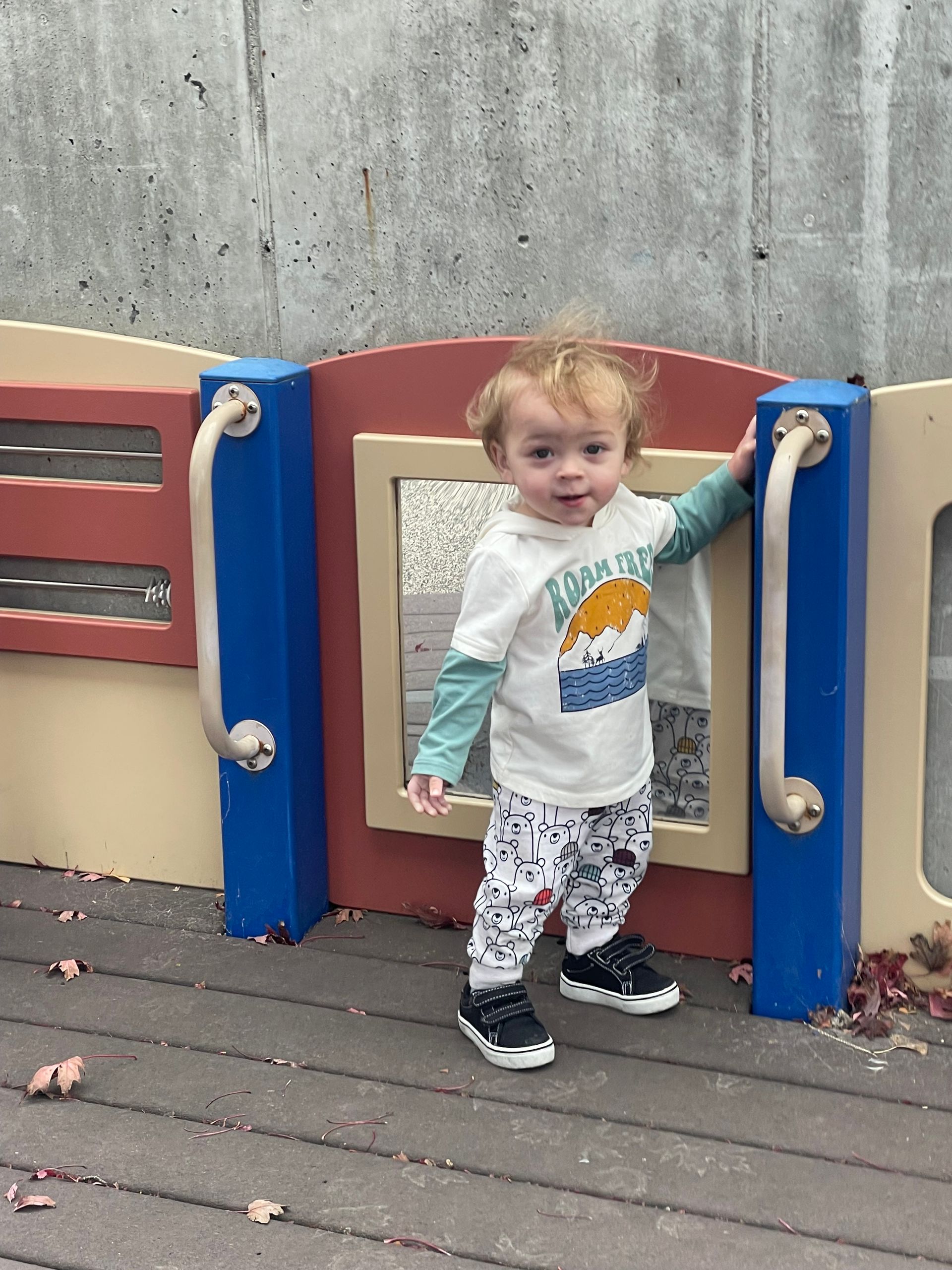 A small child stands on a playground platform, leaning against a colorful wall with cutouts, wearing a graphic tee and pants.