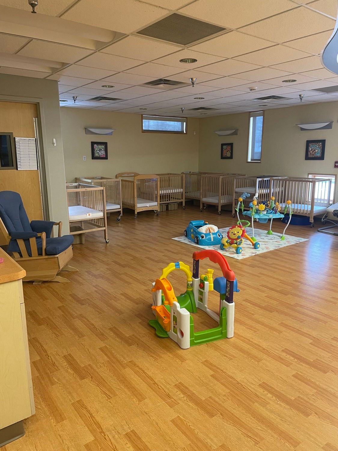 Daycare room with multiple wooden cribs, a blue rocking chair, and colorful infant floor toys on a light wood floor.