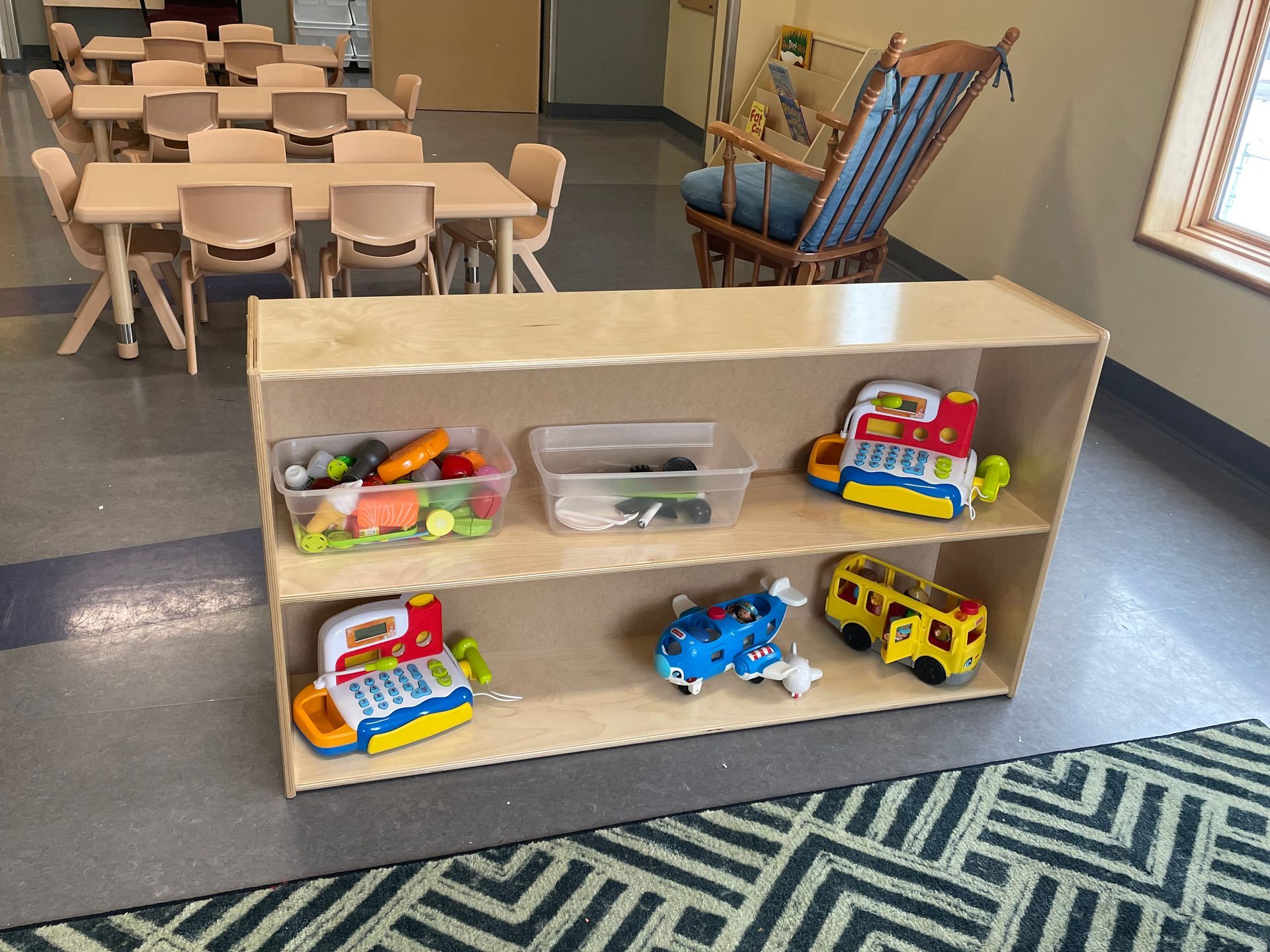 A two-tiered wooden toy shelf in a classroom, featuring storage bins and various plastic toy vehicles on display.