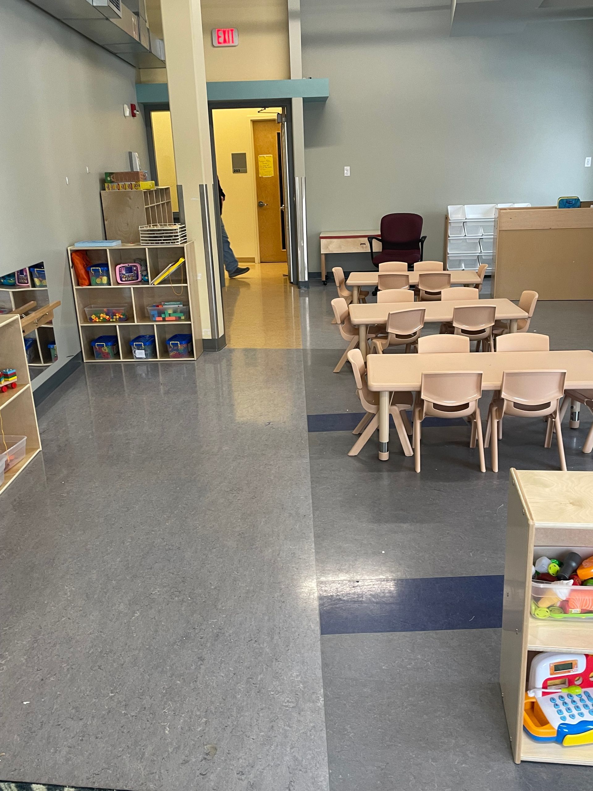 A classroom features several tables with chairs, shelves stocked with toys, and a doorway leading to another room.