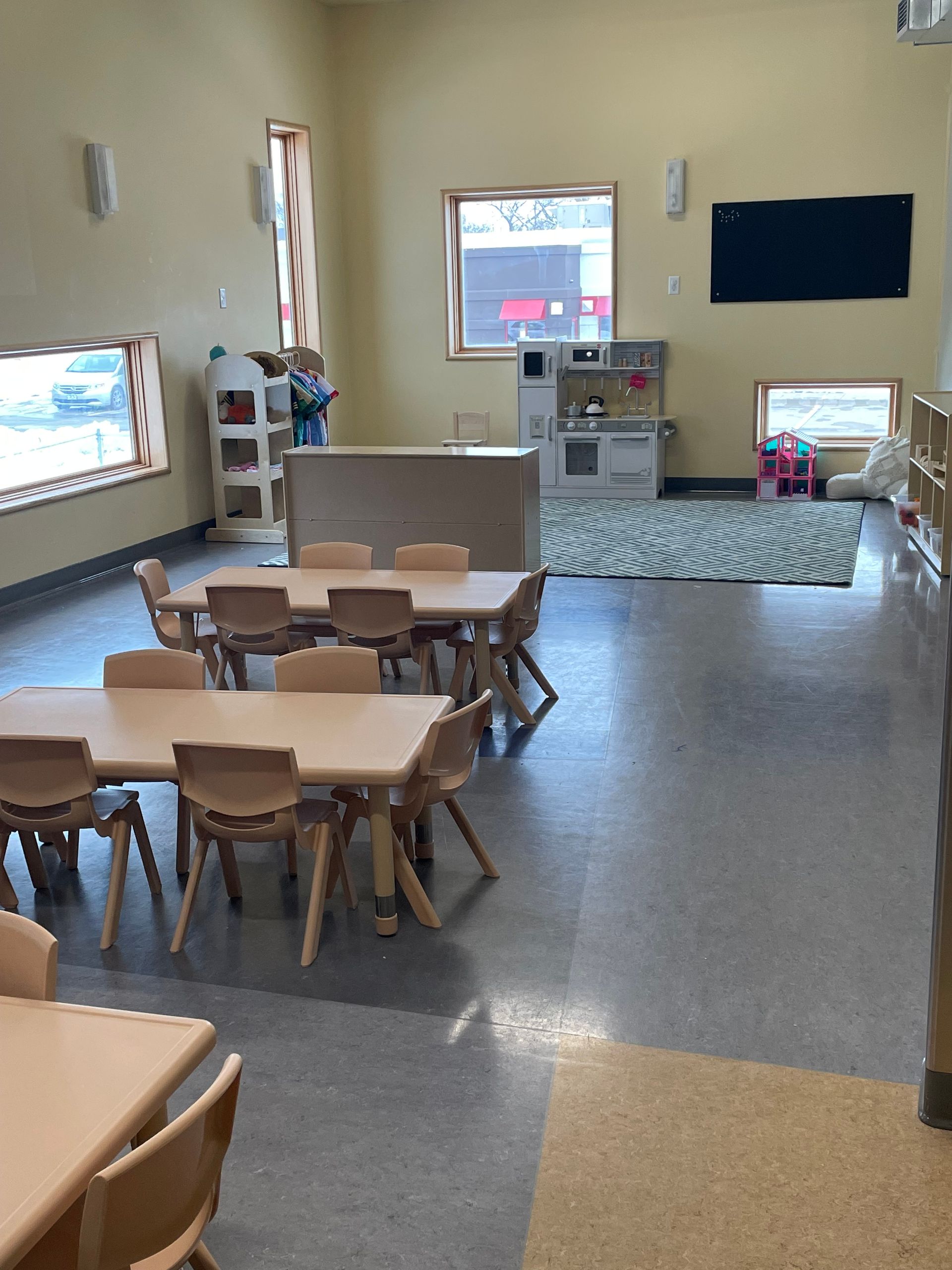 An empty preschool classroom with several small tables, chairs, and a play kitchen area on a patterned rug.
