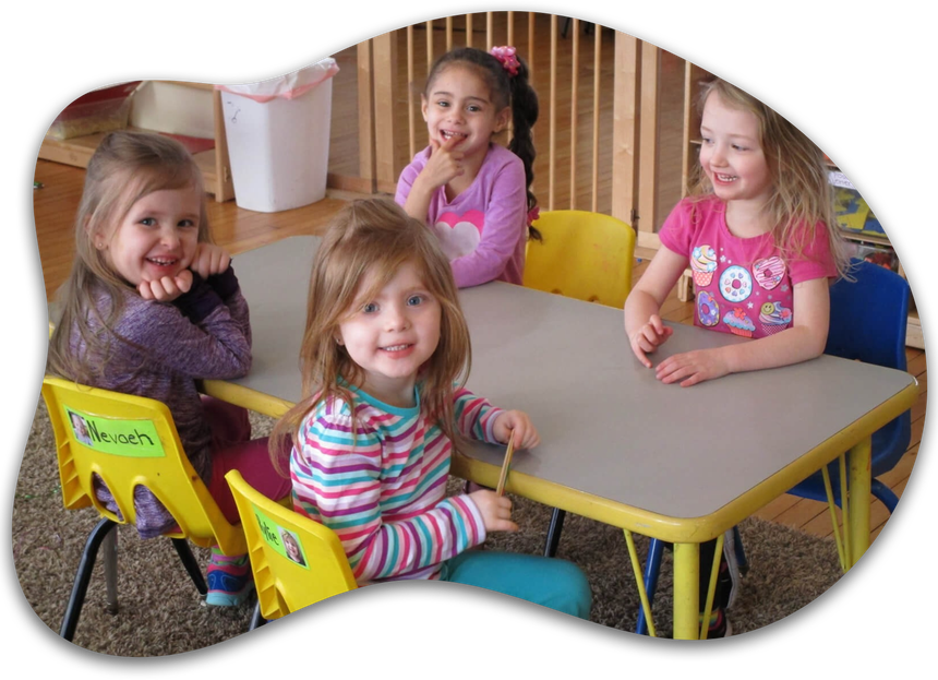 Four children sit around a table in a classroom, smiling at the camera.