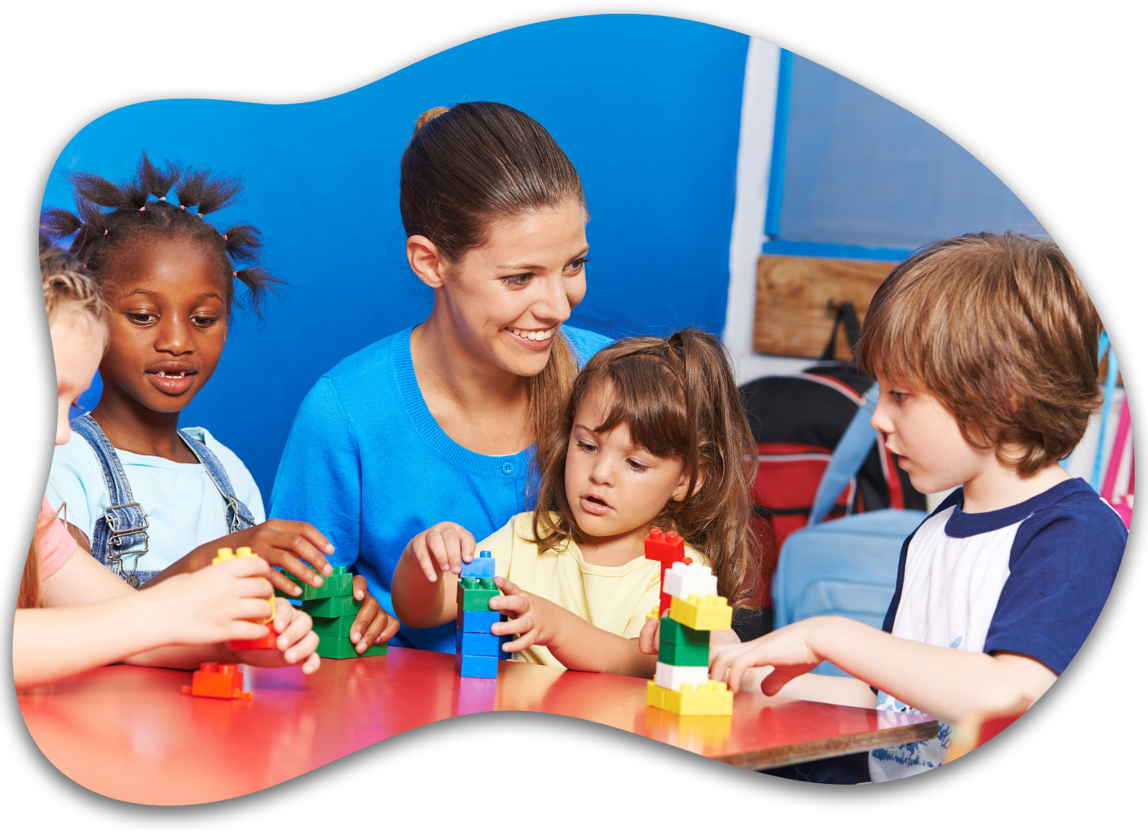 A teacher and four children sit at a red table, smiling as they build structures with colorful plastic blocks.