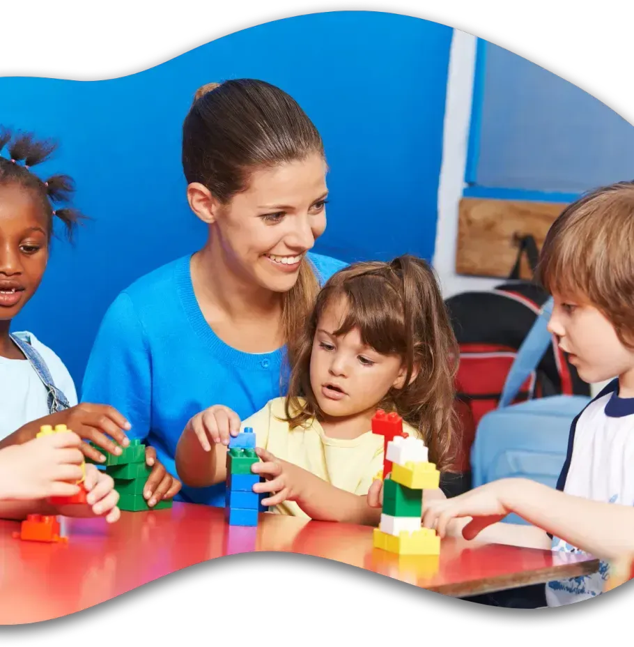 A teacher and children sit at a red table, smiling as they build structures with colorful plastic building blocks.