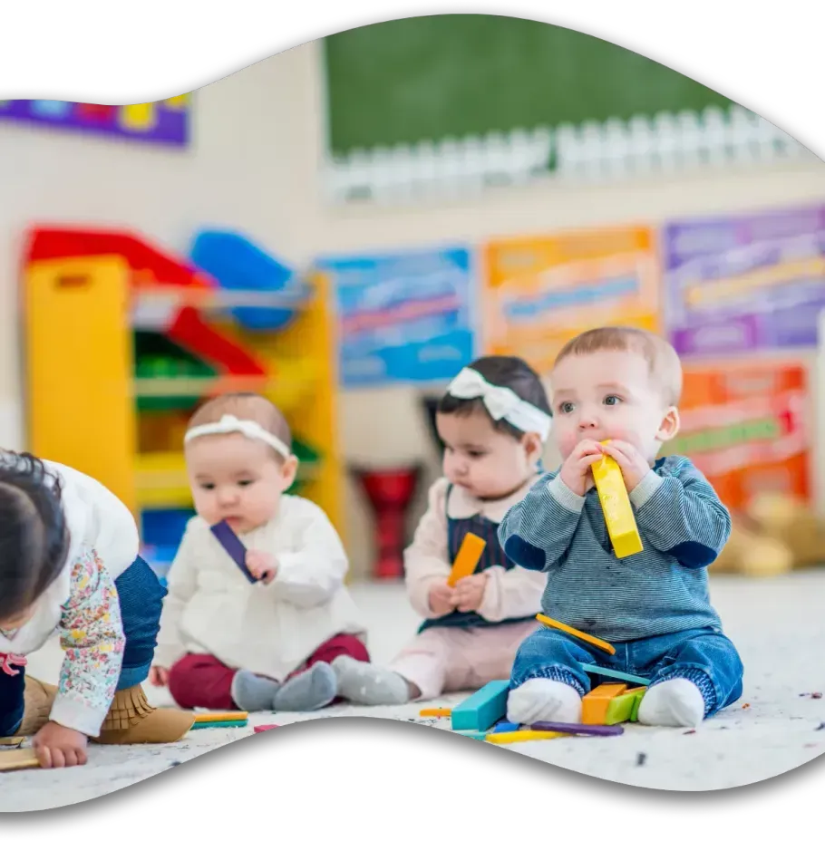 Three infants sit on a floor in a colorful classroom, playing with assorted plastic building blocks.