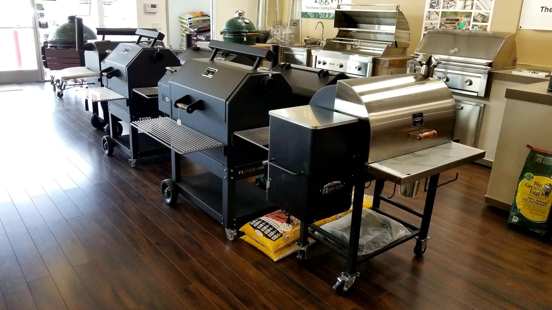 A showroom displays various black and stainless steel grills. A bag of charcoal sits nearby.