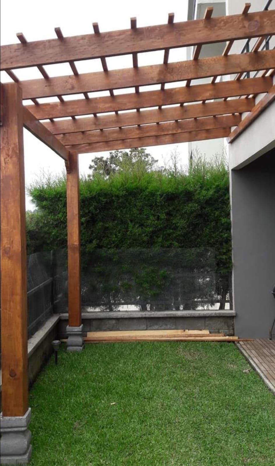 Pequeño patio trasero con pérgola de madera, césped verde y valla bordeada de setos.