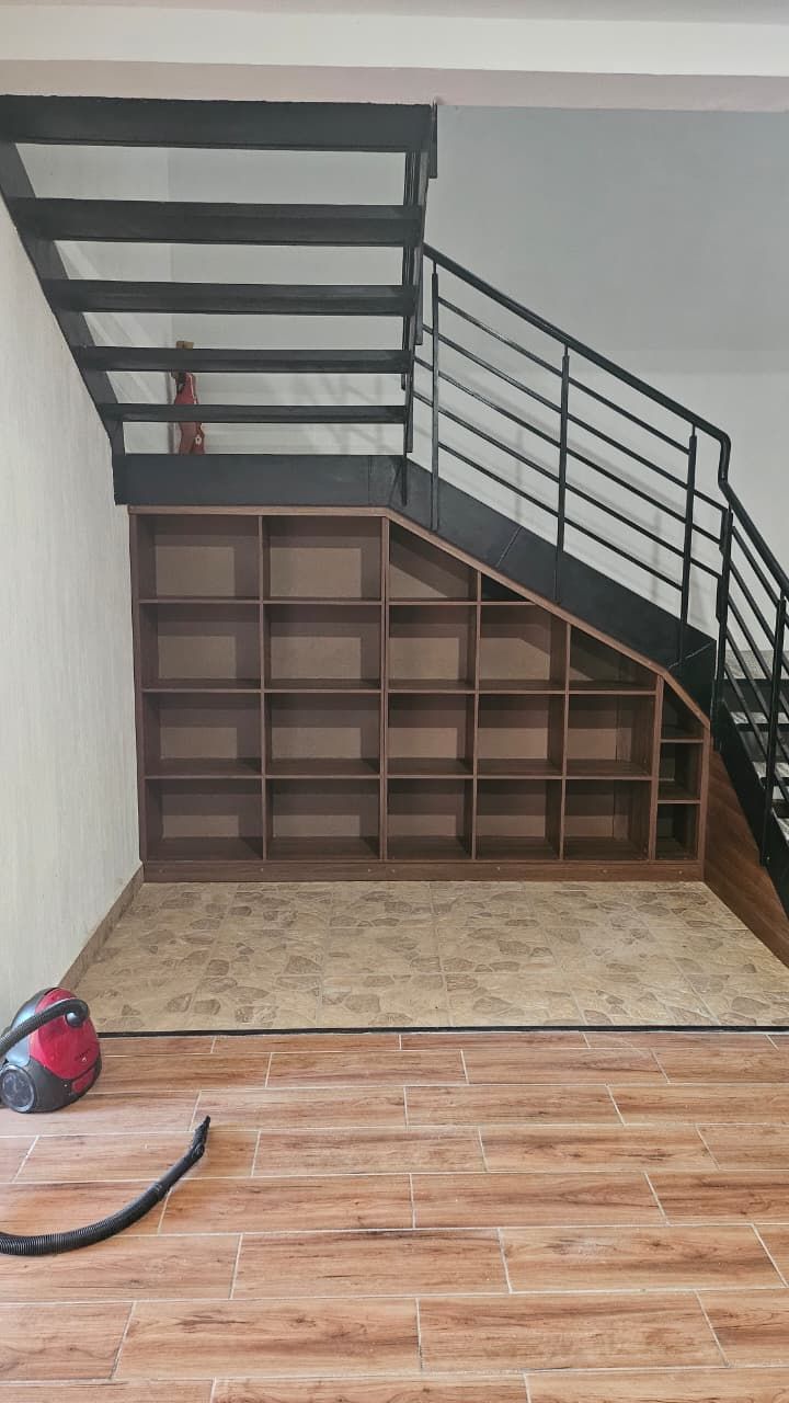 Escalera interior con barandilla oscura sobre una pared con armarios de madera y suelo de baldosas; bolsa roja en el suelo.