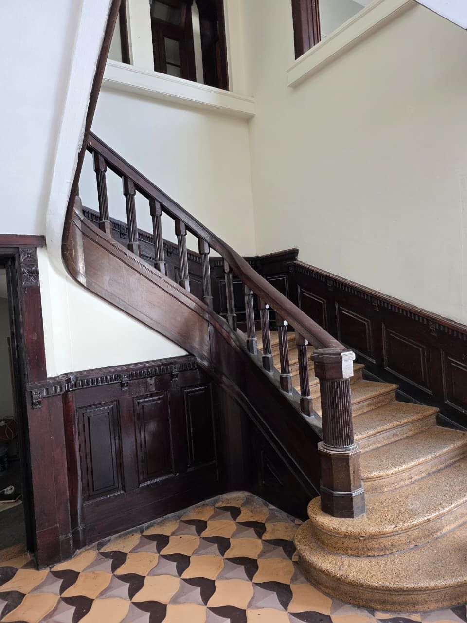 Escalera curva de madera en un interior histórico con suelo de baldosas estampadas y paneles de madera oscura.
