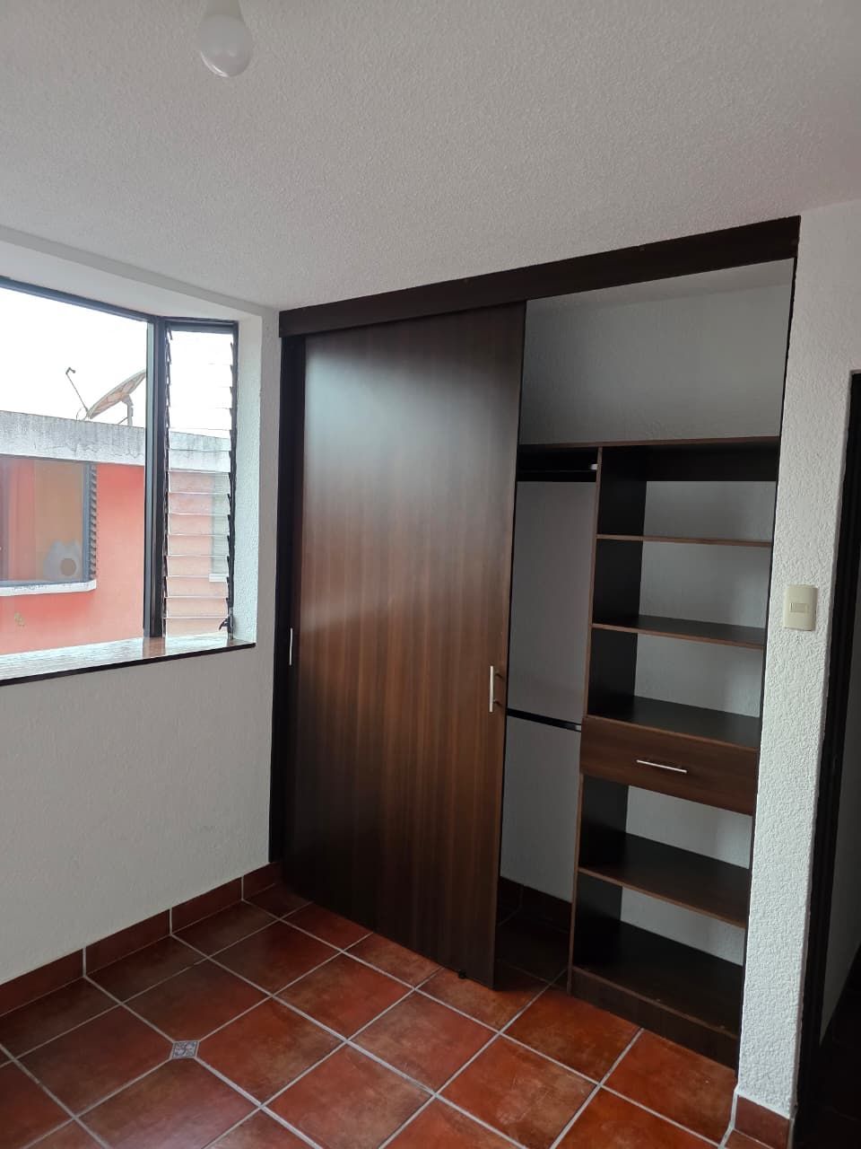 Habitación vacía con suelo de baldosas de terracota, ventana abierta y un armario de madera oscura con puerta corredera.