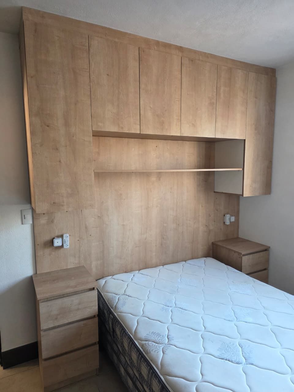 Habitación pequeña con cabecero de madera con estantes empotrados, cama blanca y dos mesitas de noche.