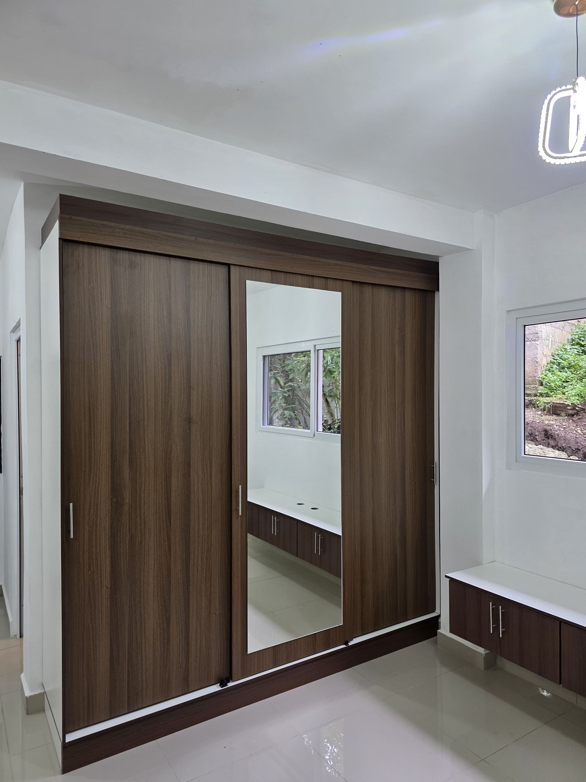 Dormitorio con armario de puertas correderas de madera y espejo, paredes blancas y un banco junto a la ventana.