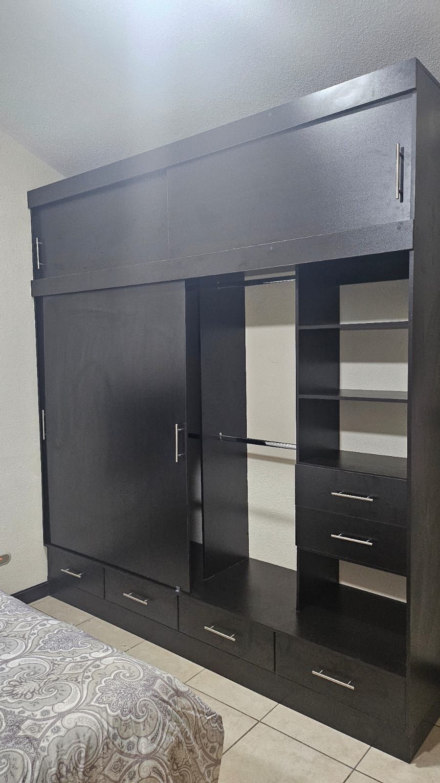 Armario negro con puertas correderas, estantes y cajones en un dormitorio, contra una pared clara.