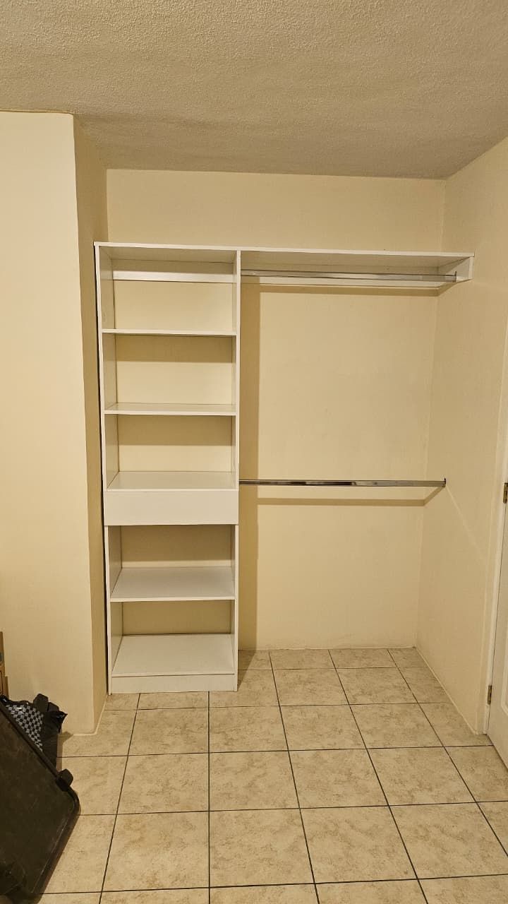 Armario vacío con estantes y barras para colgar ropa blancas, paredes beige y suelo de baldosas.