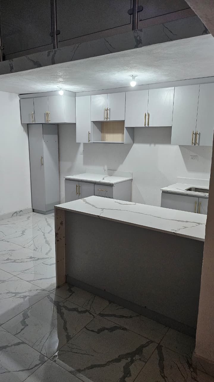 Cocina moderna blanca con isla, armarios y suelo con estampado de mármol bajo luces de techo.