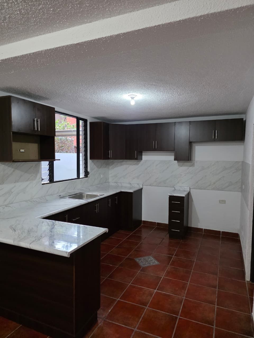 Cocina pequeña con armarios oscuros, encimeras blancas con aspecto de mármol y suelo de baldosas rojas.