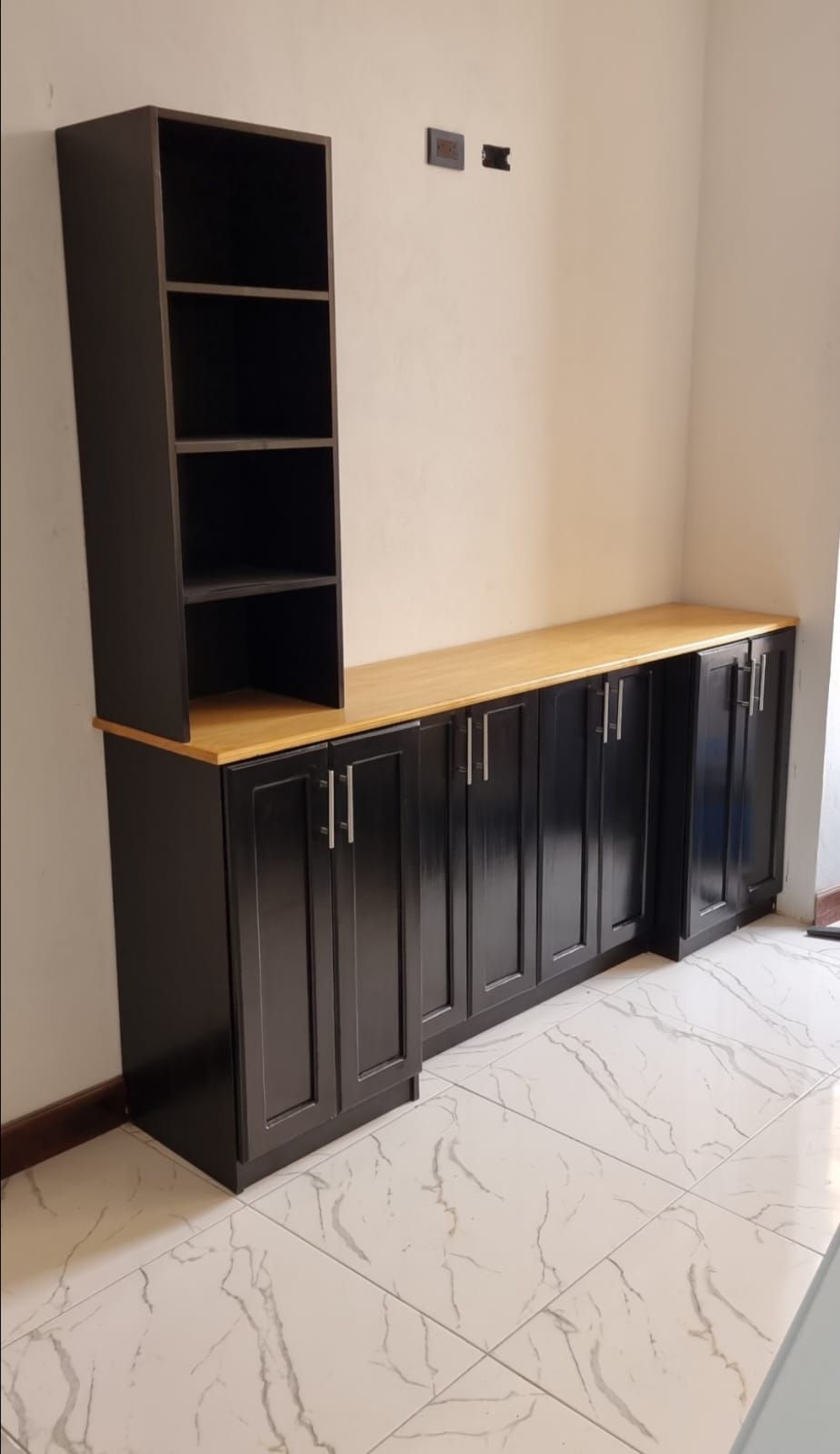 Armario negro con encimera de madera y estantería alta contra una pared beige en una habitación con suelo de baldosas.