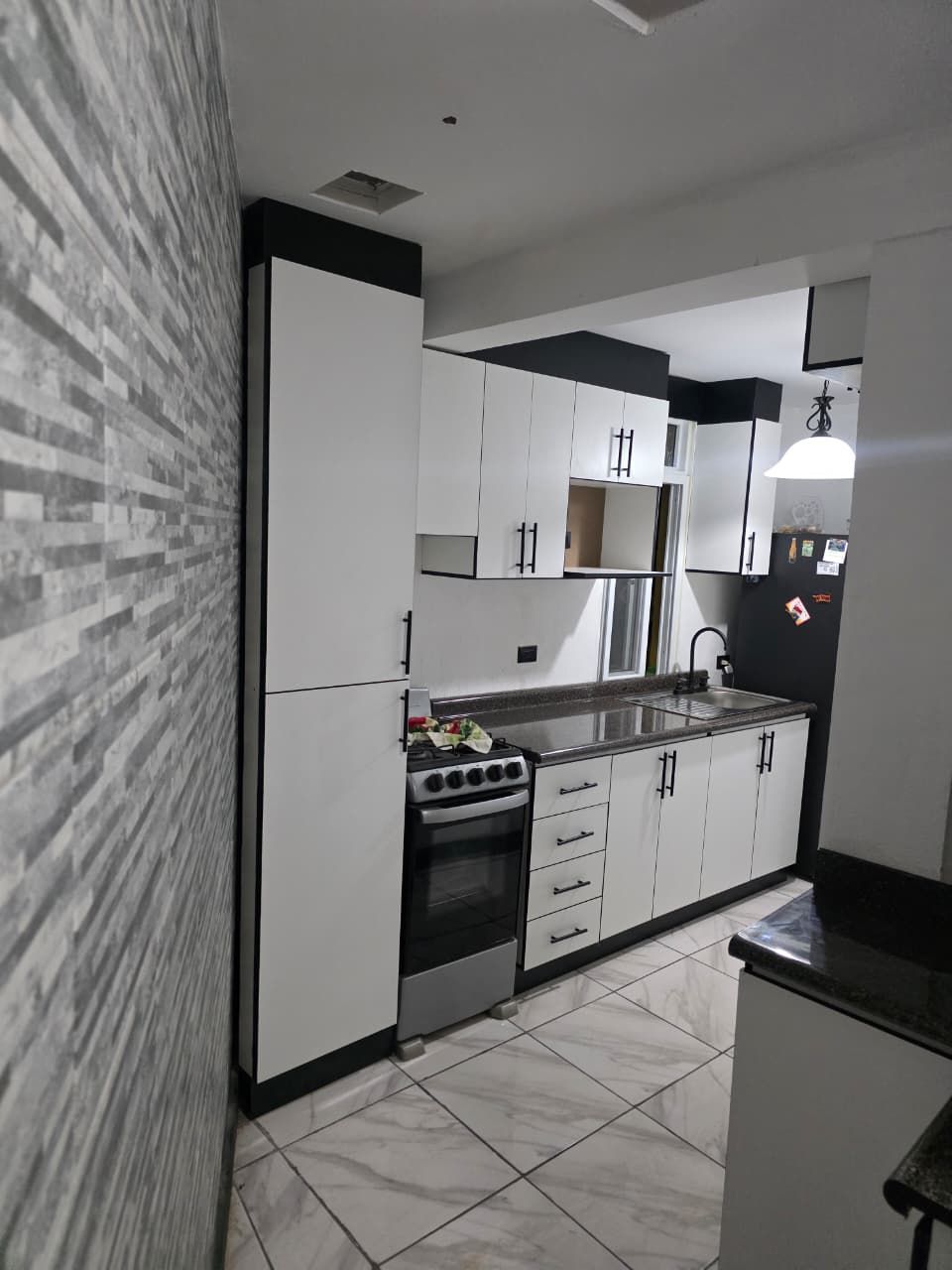 Cocina estrecha con armarios blancos, encimeras negras, suelo de baldosas y electrodomésticos de acero inoxidable.