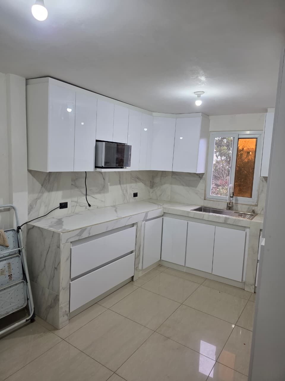 Cocina moderna de color blanco brillante con armarios, suelo de baldosas y una ventana sobre el fregadero.