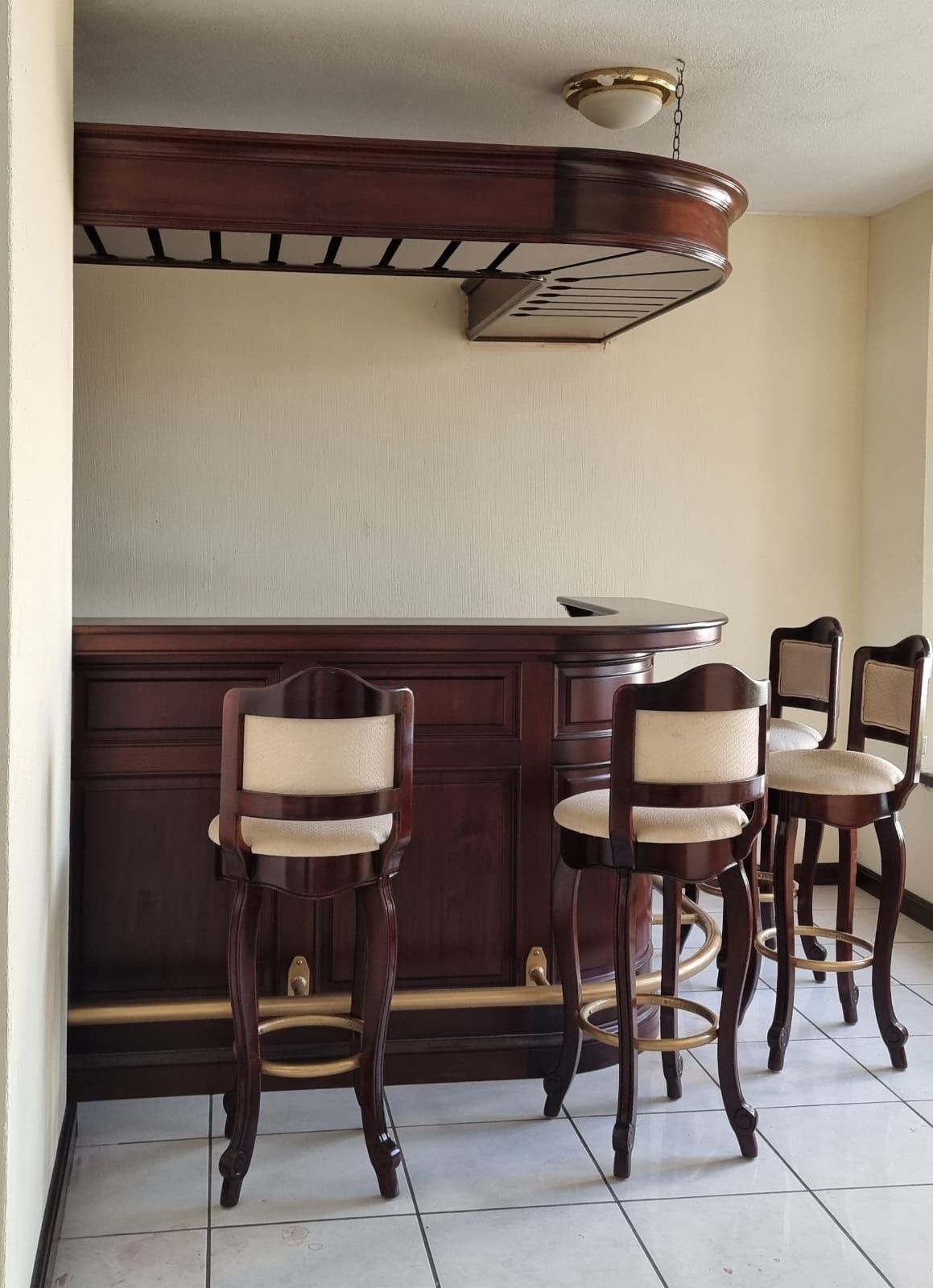 Zona de bar en casa con tres taburetes altos de madera, armarios de madera oscura y una estantería colgante en el techo.