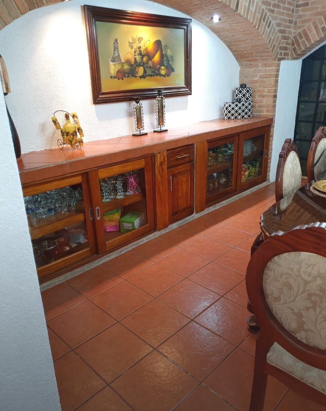 Aparador de madera con puertas de cristal contra paredes de ladrillo, con lámpara encima, cuadros enmarcados