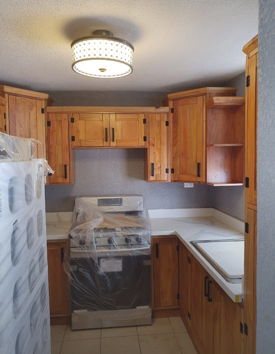 Cocina pequeña con armarios de madera, encimeras blancas y una estufa cubierta de plástico.