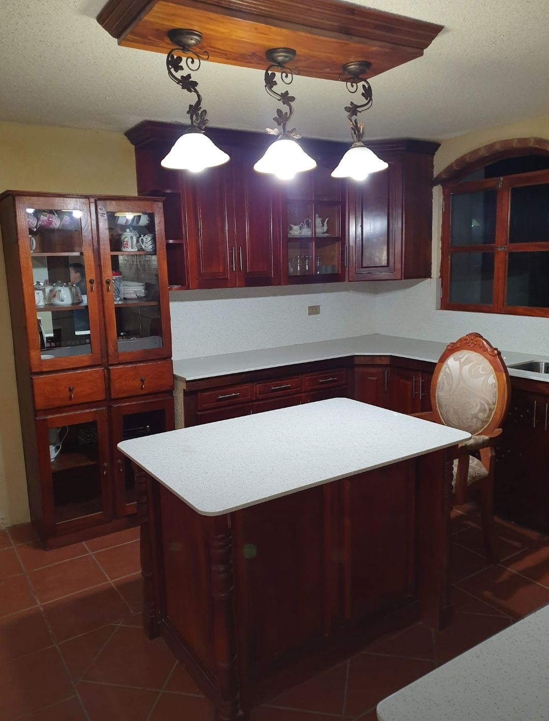 Cocina moderna con gabinetes de madera oscura, encimera blanca en la isla, lámparas colgantes y piso de baldosas.