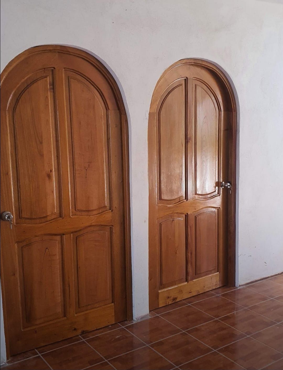 Dos puertas arqueadas de madera sobre un suelo de baldosas, junto a una pared blanca.