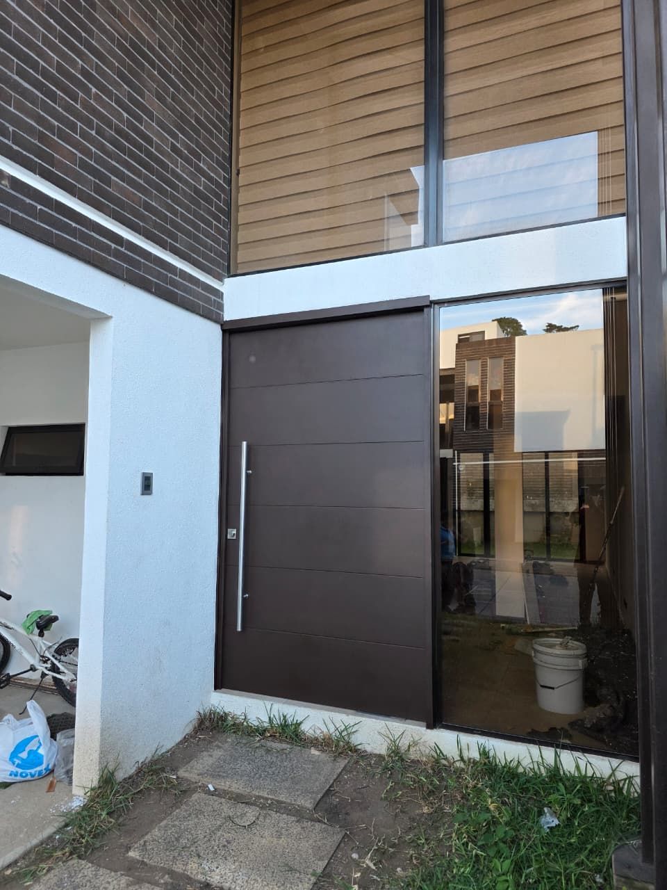 Entrada a una casa moderna con puerta principal oscura, panel lateral de vidrio y un pequeño porche pavimentado con plantas.
