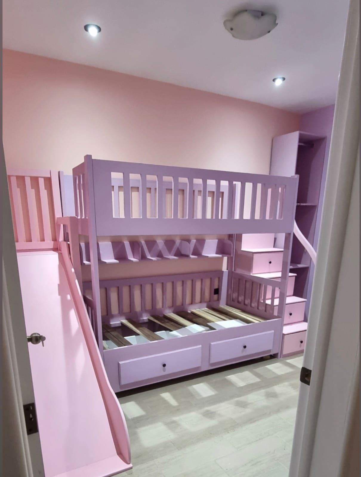 Habitación infantil rosa con una litera morada y un tobogán, vista a través de una puerta.