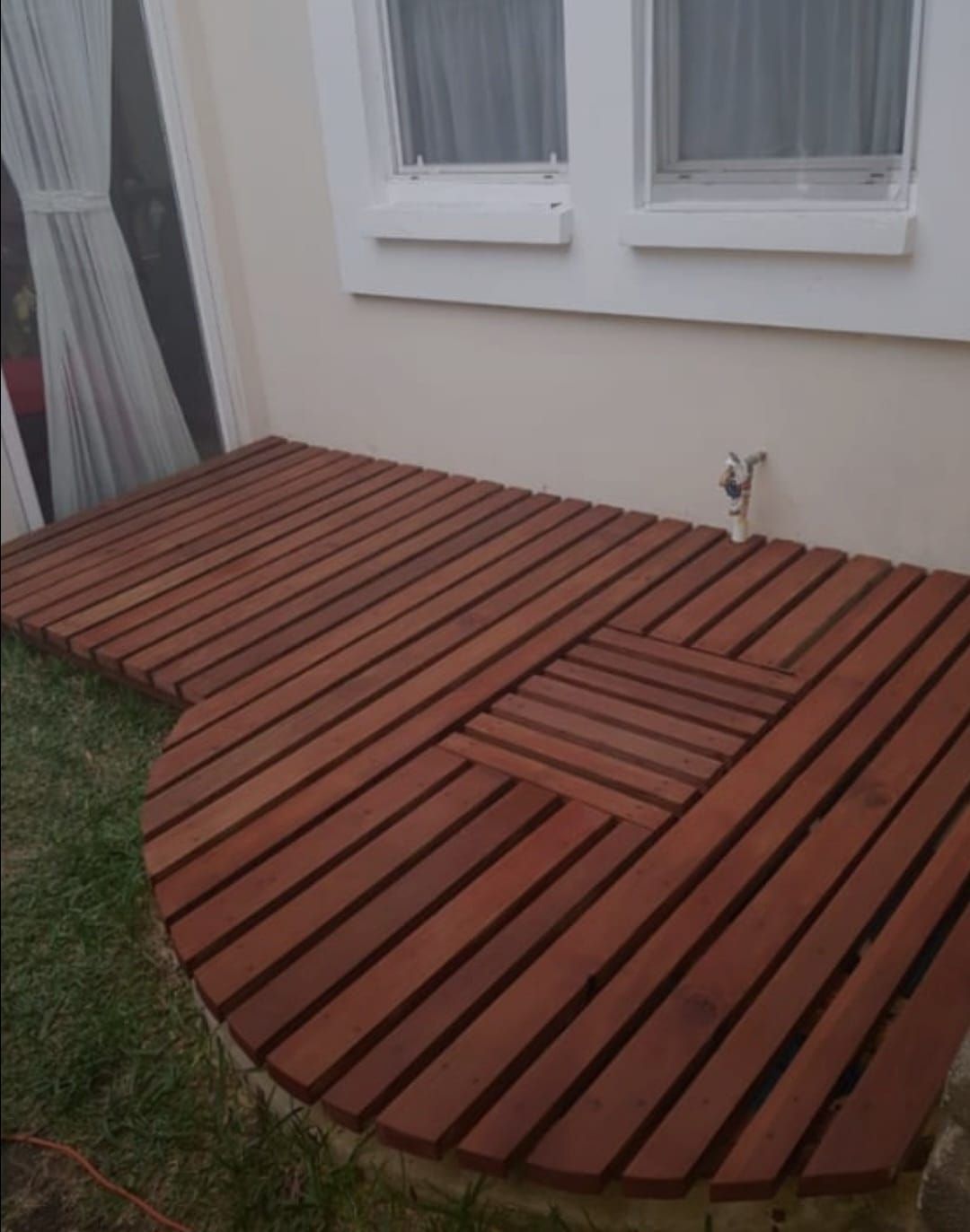 Terraza de madera con listones junto a la pared y las ventanas de la casa, con un borde curvo y un pequeño borde de césped.