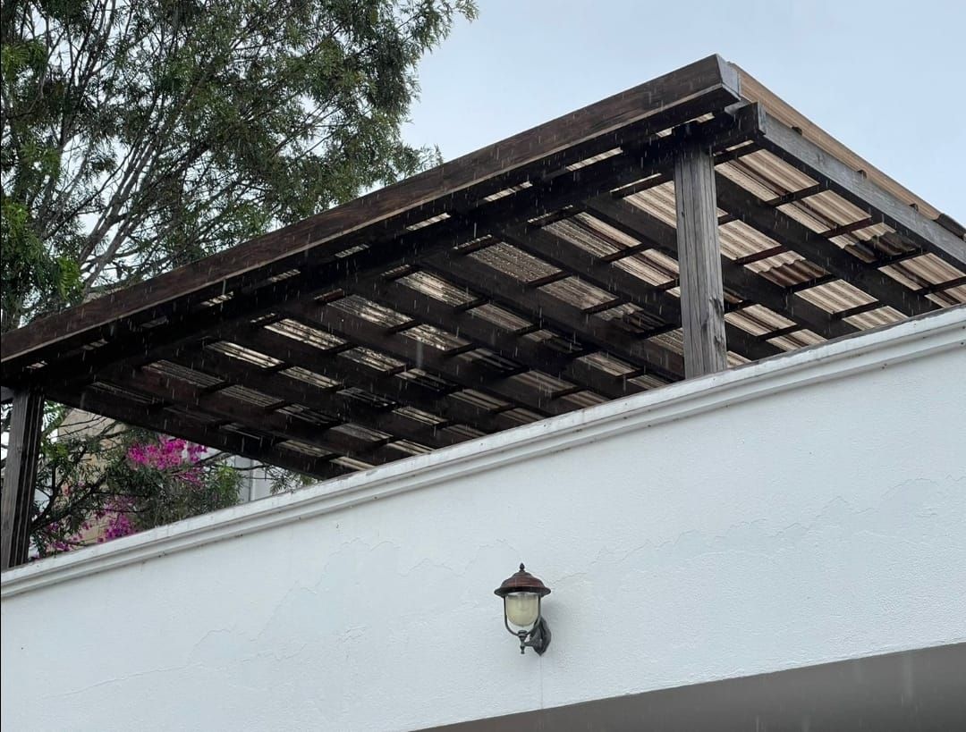 Pared blanca con una linterna debajo de un techo de pérgola de madera inclinado y árboles al fondo.