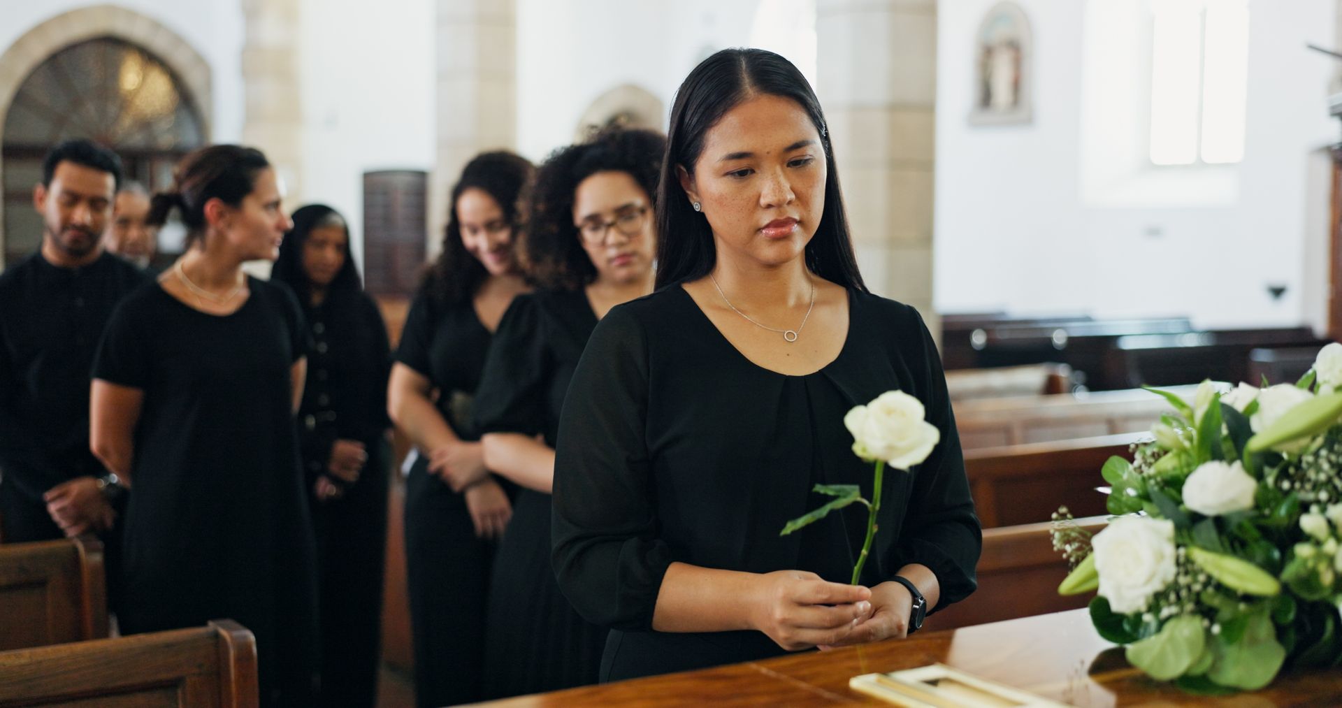 Mourners in black stand in a church as a woman holds a white rose near a coffin with flowers