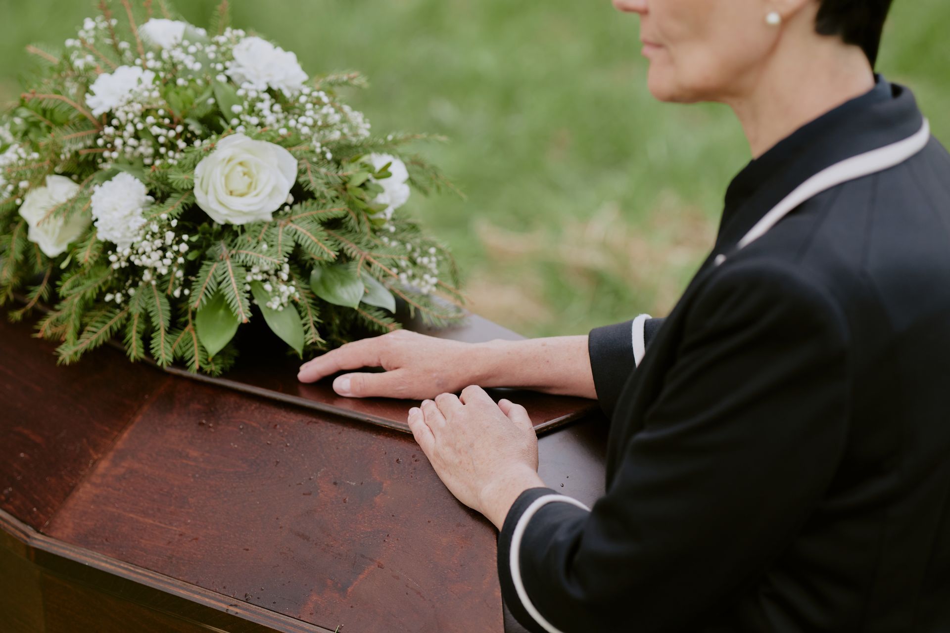 Person in black jacket resting hands on a coffin with white flower arrangement nearby