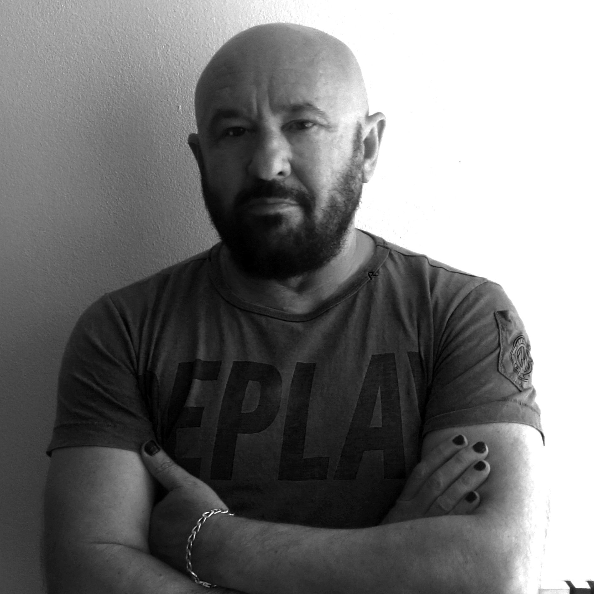 Artist Jose Gomez with beard, arms crossed, wearing a t-shirt. Black nail polish, stern expression.