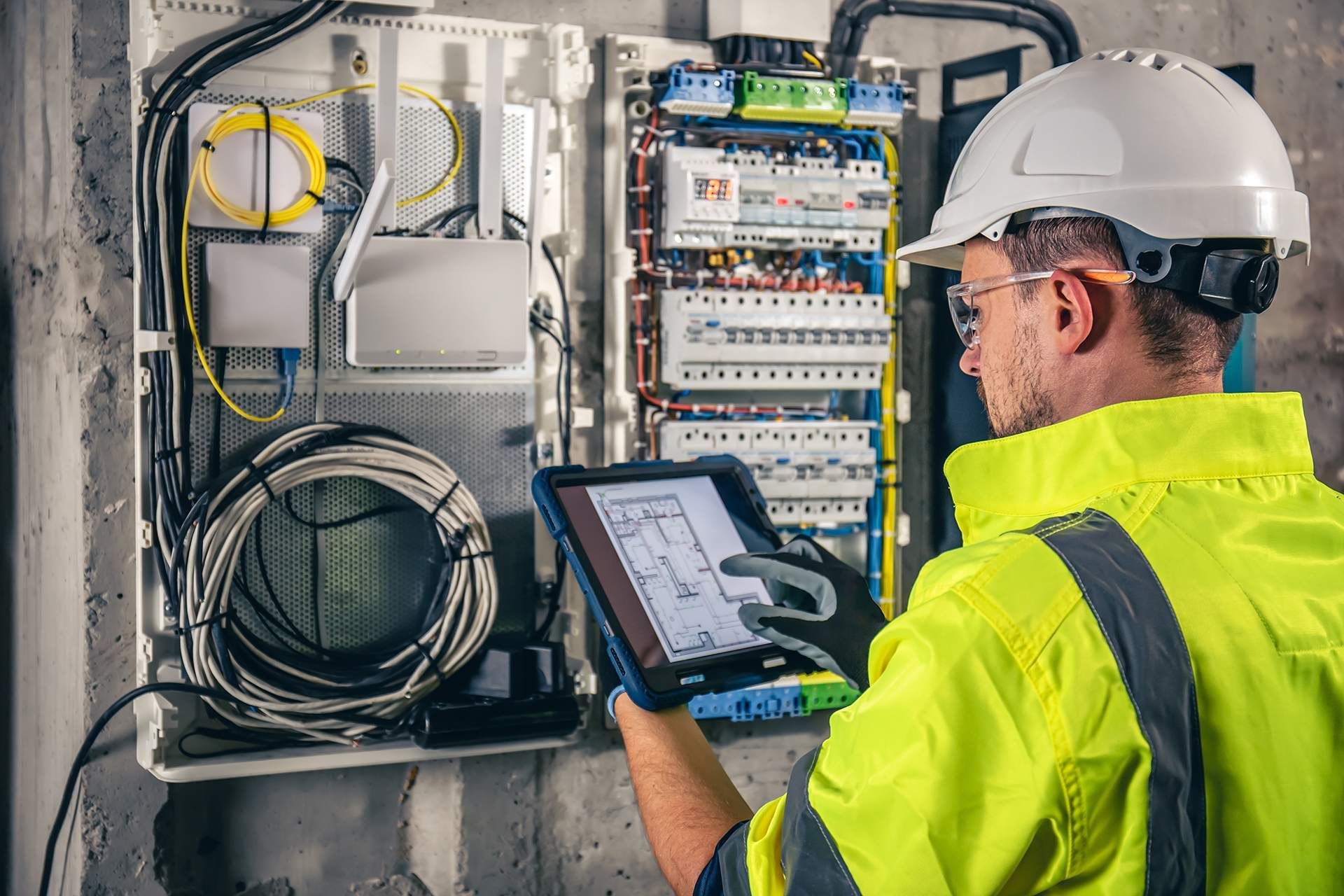 Electrician in a hard hat and safety vest examining a circuit box with a tablet.