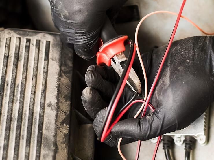 Hands in black gloves using a multimeter to test wires in an engine bay.