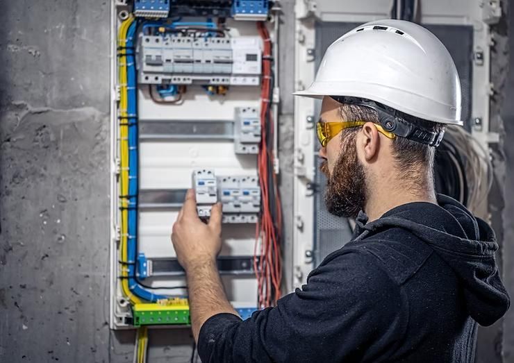 Electrician in a hard hat and safety glasses examines electrical panel.