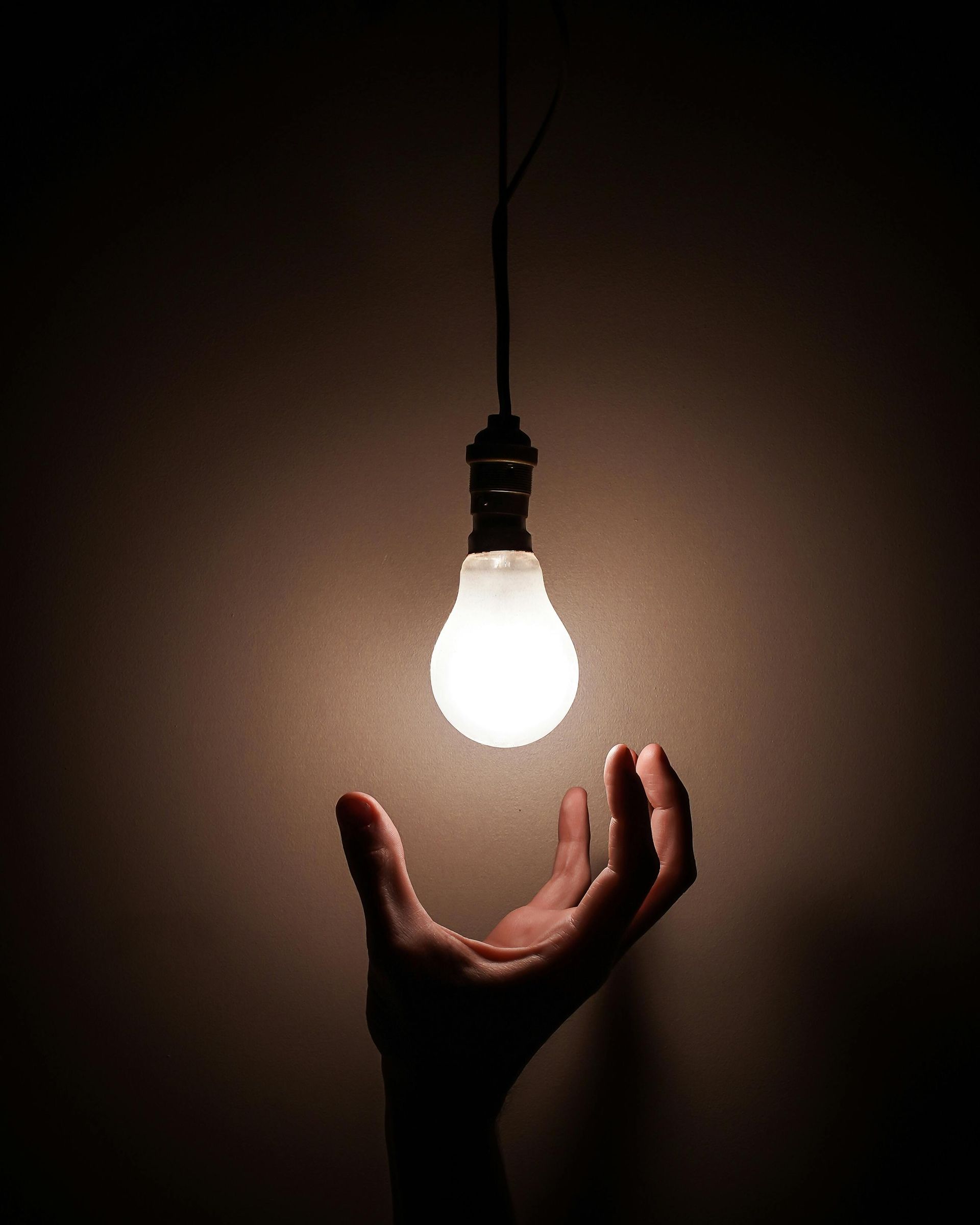 Hand reaching towards a lit lightbulb suspended in a dark space.