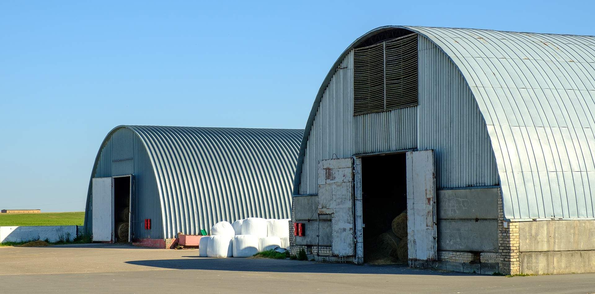 Two arched metal buildings on a paved surface with open doors against a clear blue sky.