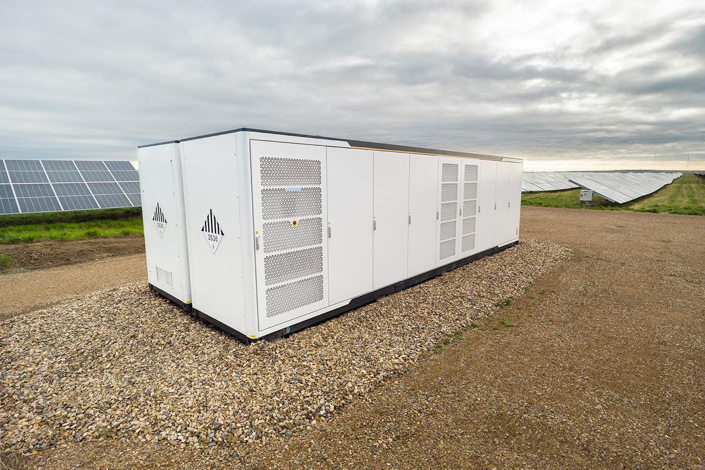 White energy storage unit with ventilation panels, gravel base, solar panels in background.
