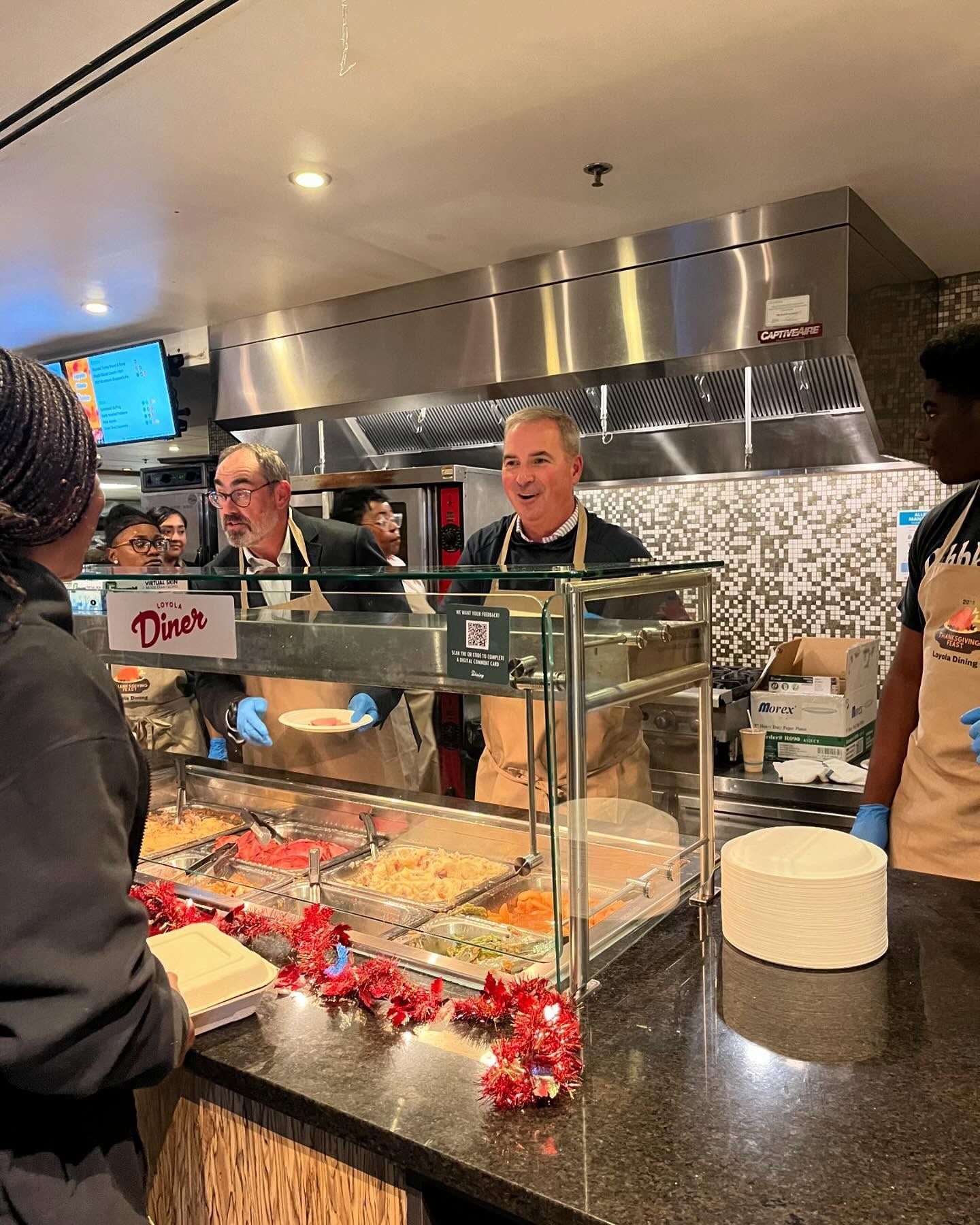 People behind a deli counter serving food in a buffet-style kitchen with stainless steel and glass display.