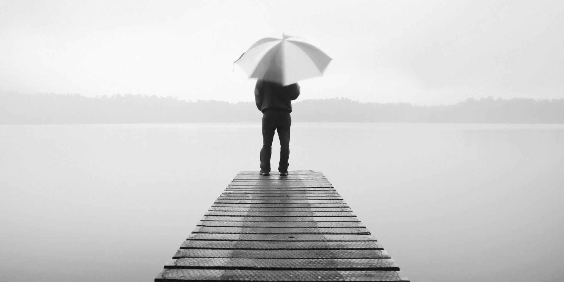 Person holding an umbrella stands on a wooden dock in a foggy lake scene.
