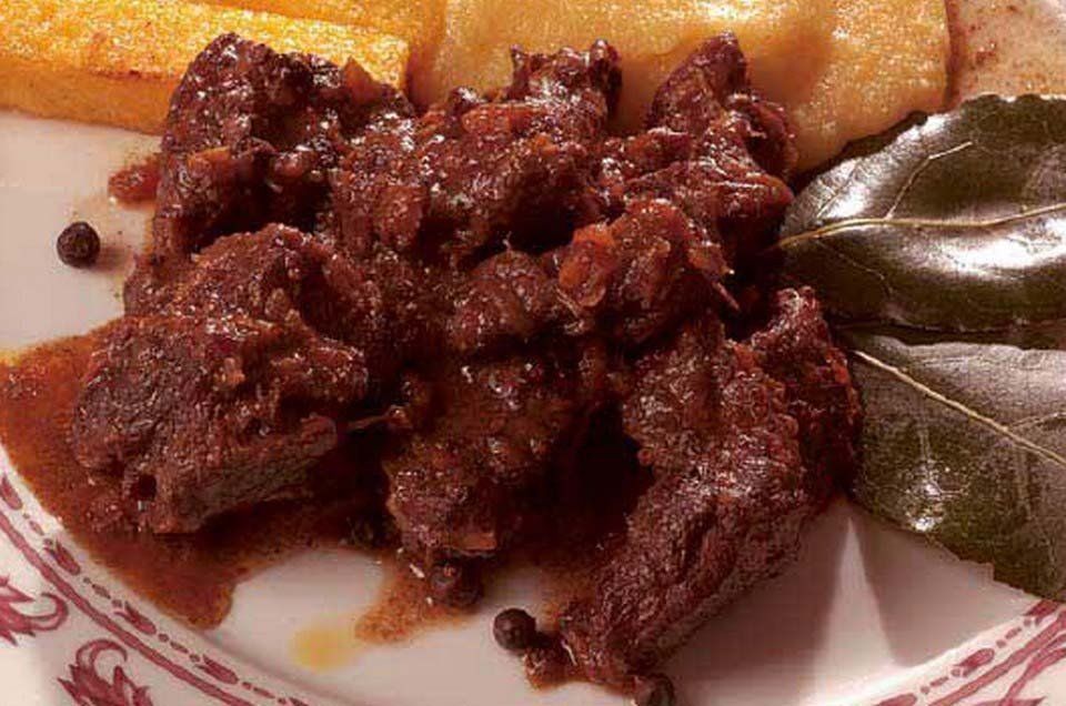 Carne cotta di colore rosso-marrone scuro su un piatto bianco con un contorno giallo e foglie verdi.